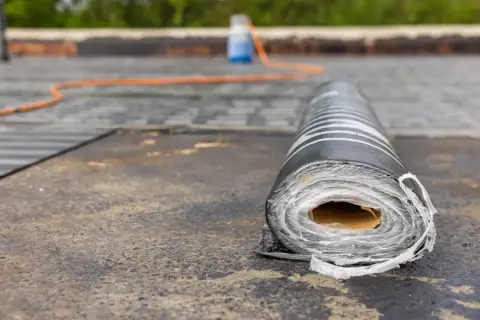 Rolka papy na dachu, gotowa do pracy. W tle pomarańczowy wąż i butla z płynem, wskazujące na proces, jak kłaść papę.