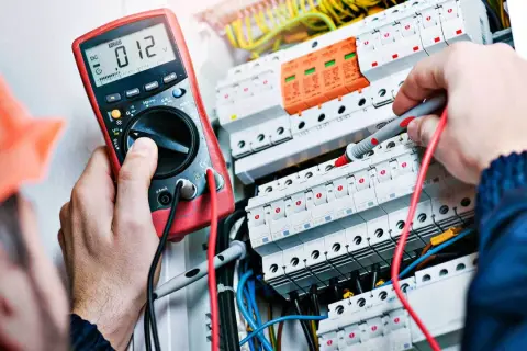 Elektryk w pomarańczowym kasku mierzy napięcie multimetrem w skrzynce z bezpiecznikami.