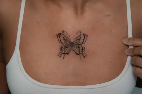 Zartes Fineline-Tattoo eines Schmetterlings zwischen den Brüsten, das wie ein filigranes Kunstwerk wirkt.