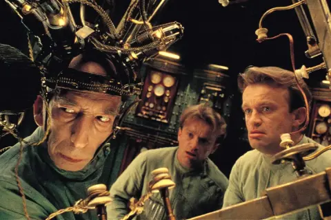 Trzej mężczyźni w dziwnych hełmach, jak z **starych filmów science fiction**, pracują przy skomplikowanej maszynie.