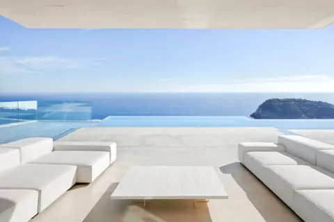 Terrasse moderne avec canapés blancs et table basse, bordée par une piscine à débordement et la mer bleue. Le **béton ciré autour de la piscine** crée une ambiance épurée et luxueuse.