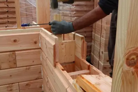 Construcción de una cabaña con madera aislante térmico. Un trabajador martilla una pieza de madera para encajarla.