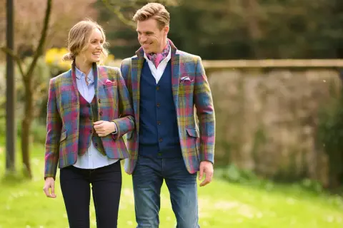 Ein Paar in farbenfrohen Tweed-Jacken und Westen spaziert durch einen sonnigen Park. Was ist Tweed? Ein robuster, traditioneller Stoff.
