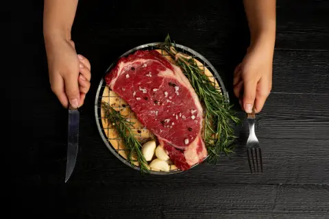 Ojo de bife crudo, sazonado con sal y pimienta, con romero y ajos, listo para cocinar. Manos sostienen cuchillo y tenedor.