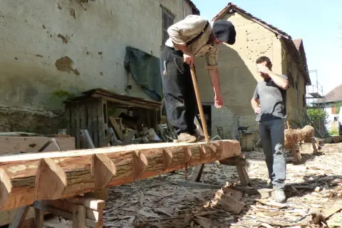 Deux hommes travaillent sur un tronc d'arbre, pratiquant l'équarrissage bois avec une hache. Le bois est façonné avec soin.