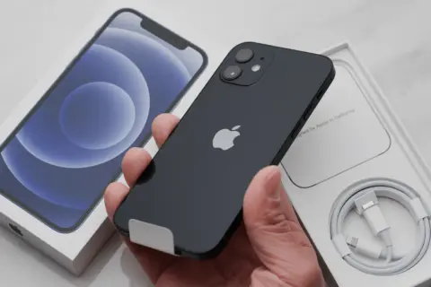Czarny iPhone 12 w ręce, obok pudełko z telefonem i kablem. Chcesz wiedzieć, jak resetować iPhone?