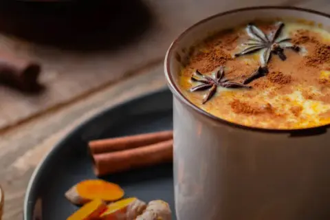 Eine Tasse goldene Milch, garniert mit Sternanis und Zimt. Daneben Zutaten für das goldene Milch Rezept: Kurkuma, Ingwer, Zimtstangen und Honig.
