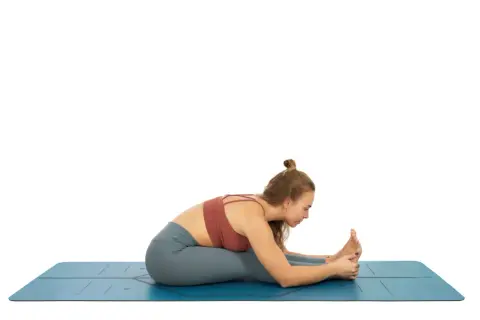 Mujer en esterilla azul, practicando una de las variantes de uttanasana, con las piernas estiradas y el torso inclinado hacia adelante, sujetando los pies.