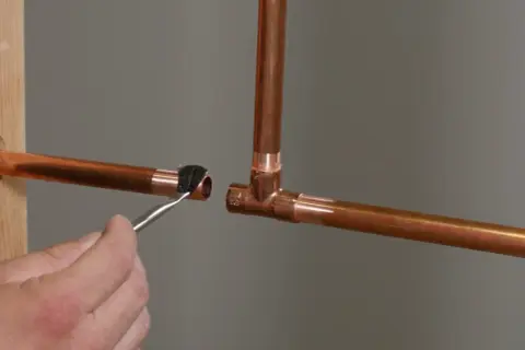 A hand applies flux with a brush to a copper pipe end, preparing it for soldering to a copper tee fitting.