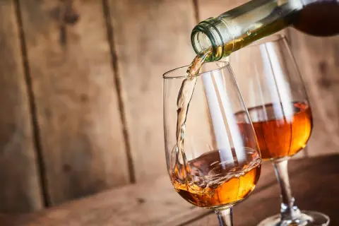 Un verre se remplit d'un vin paillé ambré, un autre verre est déjà servi. Ambiance chaleureuse sur fond de bois.