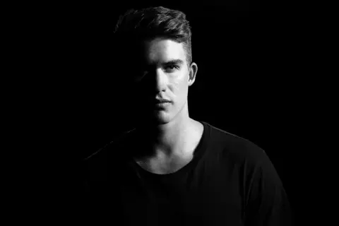 Un jeune homme au regard intense, éclairé par un côté, avec un fond noir pour mettre en valeur son visage.