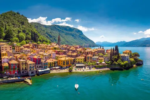 Village coloré au bord des lacs italiens, avec des montagnes verdoyantes en arrière-plan et un voilier sur l'eau turquoise.