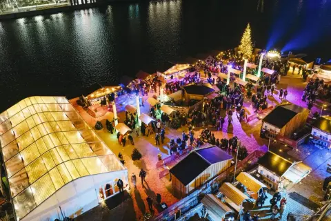 Buntes Treiben auf einem weihnachtsmarkt an der Spree. Menschen schlendern zwischen beleuchteten Buden und einem großen Weihnachtsbaum.