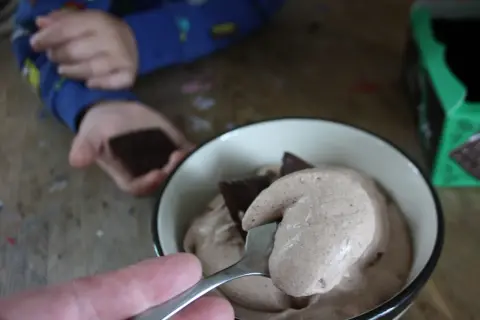 En sked med krämig after eight mousse lyfts ur en skål. En barnhand håller en chokladbit i bakgrunden.