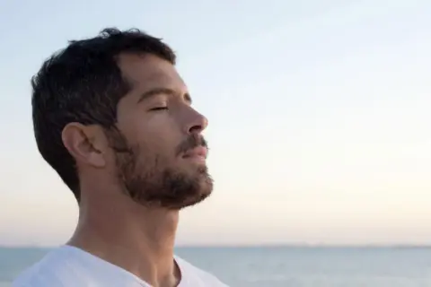 Un homme au profil serein, yeux clos, savoure l'air marin. Sa barbe de 3 jours, d'environ 3 mm, accentue son charme naturel.
