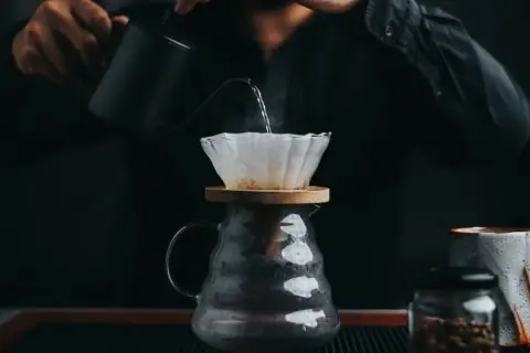En person häller varmt vatten över kaffefilter för att brygga dropp kaffe. Ånga stiger från den nybryggda drycken.