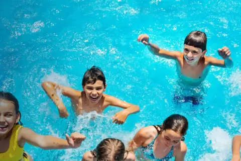 Niños riendo y chapoteando en una piscina azul, disfrutando del verano. ¡Una imagen perfecta de campamentos de inglés en España!