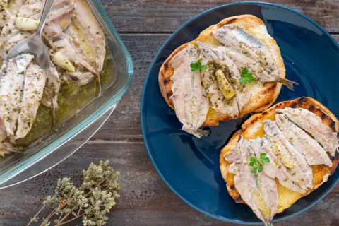 Alici al forno, profumate all'aglio e erbe, servite su crostini tostati e in una pirofila di vetro.