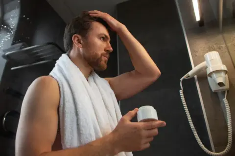 Hombre con pelo fosco se arregla el cabello con crema, envuelto en una toalla blanca.