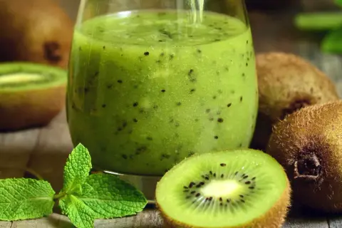 Szklanka zielonego smoothie z kiwi, pokrojone owoce kiwi i listki mięty na drewnianym tle.