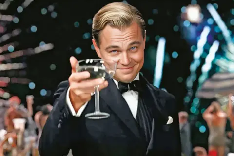 Jay Gatsby wznosi toast kieliszkiem szampana, otoczony blaskiem imprezy. Jego spojrzenie pełne jest obietnicy i tajemnicy, jak w filmie "Wielki Gatsby".