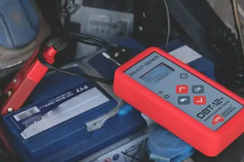 Czerwony tester akumulatorów podłączony do akumulatora samochodowego. Wyświetla 14.25V, sugerując, że problemem nie jest rozładowany akumulator, a coś innego.
