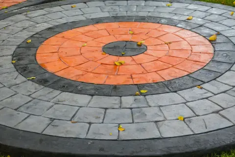 Kreisrunder Weg aus grau und orange gestrichenen Pflastersteinen, mit einigen Herbstblättern darauf.