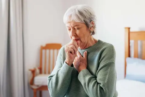 Ältere Frau hustet, kämpft mit Atemnot. Asthma kann zu einer Schwerbehinderung führen und den Alltag stark beeinträchtigen.