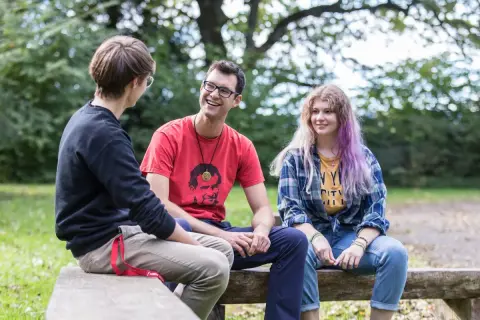 Drei junge Leute unterhalten sich lachend auf einer Bank im Freien. Der Mann in Rot trägt ein T-Shirt mit einem Porträt, das an den **Salesianer Orden** erinnert.