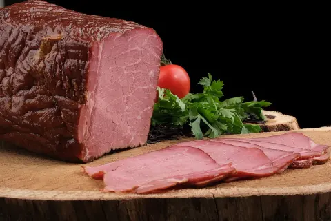 Pyszna, wędzona szynka po peklowaniu, pokrojona w cienkie plastry, z pomidorem i natką pietruszki.