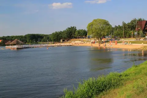 Słoneczny dzień nad jeziorem w Siemiatyczach. Plaża pełna ludzi, pomost z altaną, a w tle las i budynki. Idealne siemiatycze atrakcje.