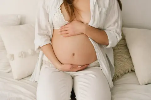 Brzuch ciężarnej opada, gdy zbliża się poród. Kobieta w białej koszuli i biustonoszu delikatnie obejmuje dłońmi swój zaokrąglony brzuch.
