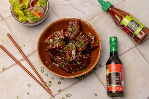 Aksamitny boczek po koreańsku w sosie, posypany sezamem i szczypiorkiem. Obok miseczka sałatki i sosy.