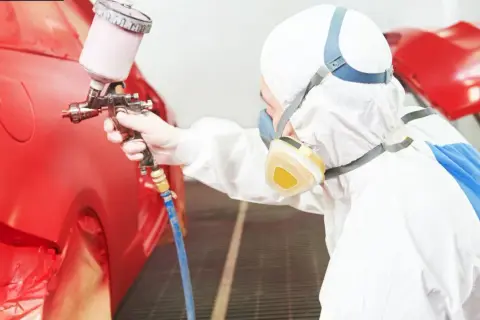 Persona in tuta protettiva e maschera effettua verniciare auto fai da te con pistola a spruzzo su un'auto rossa.