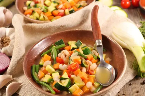 Un plato humeante de menestra de verduras, con judías verdes, zanahoria, calabacín y alubias blancas, servido con una cuchara.