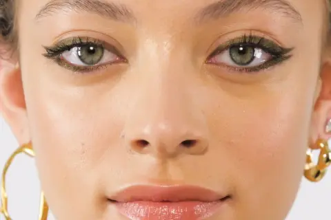 Primo piano di una ragazza con un trucco occhi audace e gioielli dorati. I suoi occhi verdi marroni sono evidenziati da un eyeliner scuro e scintillante.