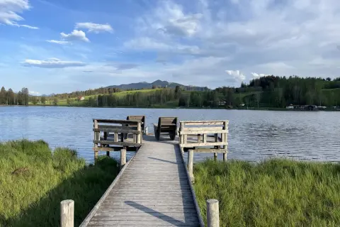 Holzsteg mit Sitzgelegenheiten am bayerischen See, umgeben von grünen Wiesen und bewaldeten Hügeln unter blauem Himmel.