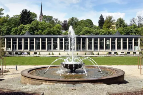 Ein Brunnen sprudelt vor einem modernen Gebäude mit Säulen und Glasfronten. Im Hintergrund ein Kirchturm und Bäume. Ein idyllischer tschechischer Kurort.