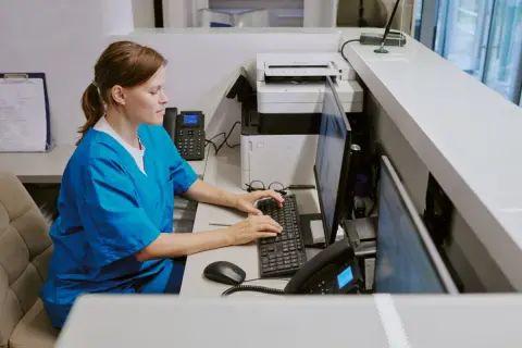 Pielęgniarka w niebieskim uniformie pracuje przy komputerze, wpisując dane. Zastanawiasz się, ile zarabia pielęgniarka?