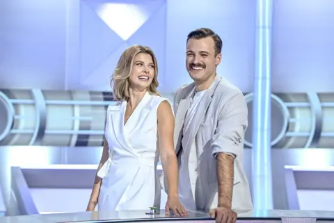 Uśmiechnięci prowadzący koło fortuny, kobieta w białej sukience i mężczyzna w jasnej marynarce, stoją przy stole w studiu.