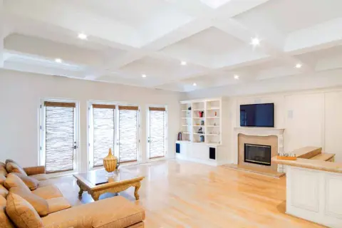 A spacious living room with a coffered ceiling featuring eight can lights. A large sectional sofa faces a fireplace and entertainment center.