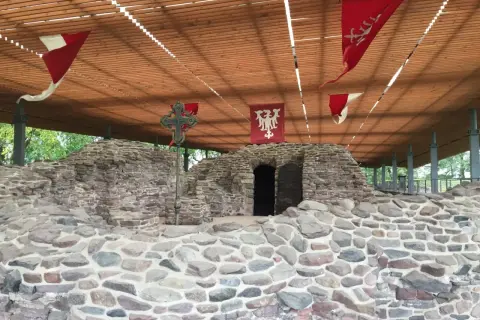 Kamienne ruiny na Ostrowiu Lednickim, z historycznymi chorągwiami i krzyżem. Wyobraź sobie, że to miejsce, skąd wyruszał dawny ostrów lednicki prom.