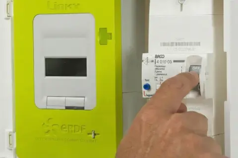Main d'un homme actionnant un disjoncteur de type BACO en position OFF, à côté d'un compteur Linky vert.