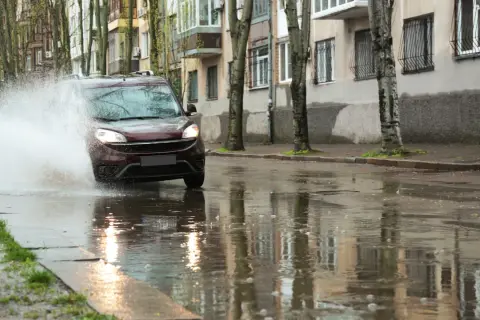 Ciemnobordowy samochód wpada w kałużę, powodując potężny rozbryzg wody. Niebezpieczny aquaplaning na mokrej ulicy.