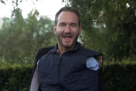 Nick Vujicic, lächelnd und mit offenem Mund, trägt eine blaue Weste über einem Hemd.