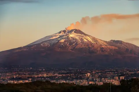 Etna, najwyższy wulkan Europy, dymi na tle zachodzącego słońca. U jego stóp rozciąga się miasto.