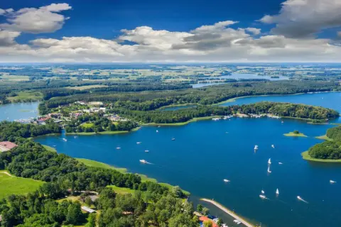 Widok z lotu ptaka na malownicze jezioro z żaglówkami. Z tamtej strony jeziora tekst widać zielone lasy i pola.