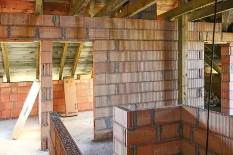 Budowa domu z bloczków z betonu komórkowego. Widać ściany z pustaków, drewniane belki stropowe i materiały budowlane, jak klej czy zaprawa do betonu komórkowego.