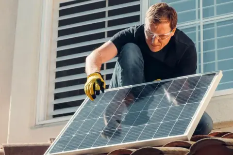 Mężczyzna w okularach ochronnych montuje panel słoneczny na dachu, walcząc ze zmianami klimatycznymi.