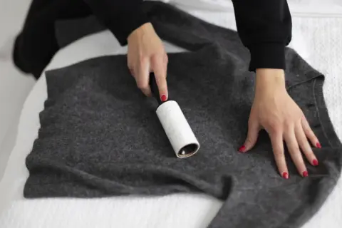 Dłonie z czerwonymi paznokciami usuwają kłaczki z szarej, zmechaconej bluzy za pomocą wałka.